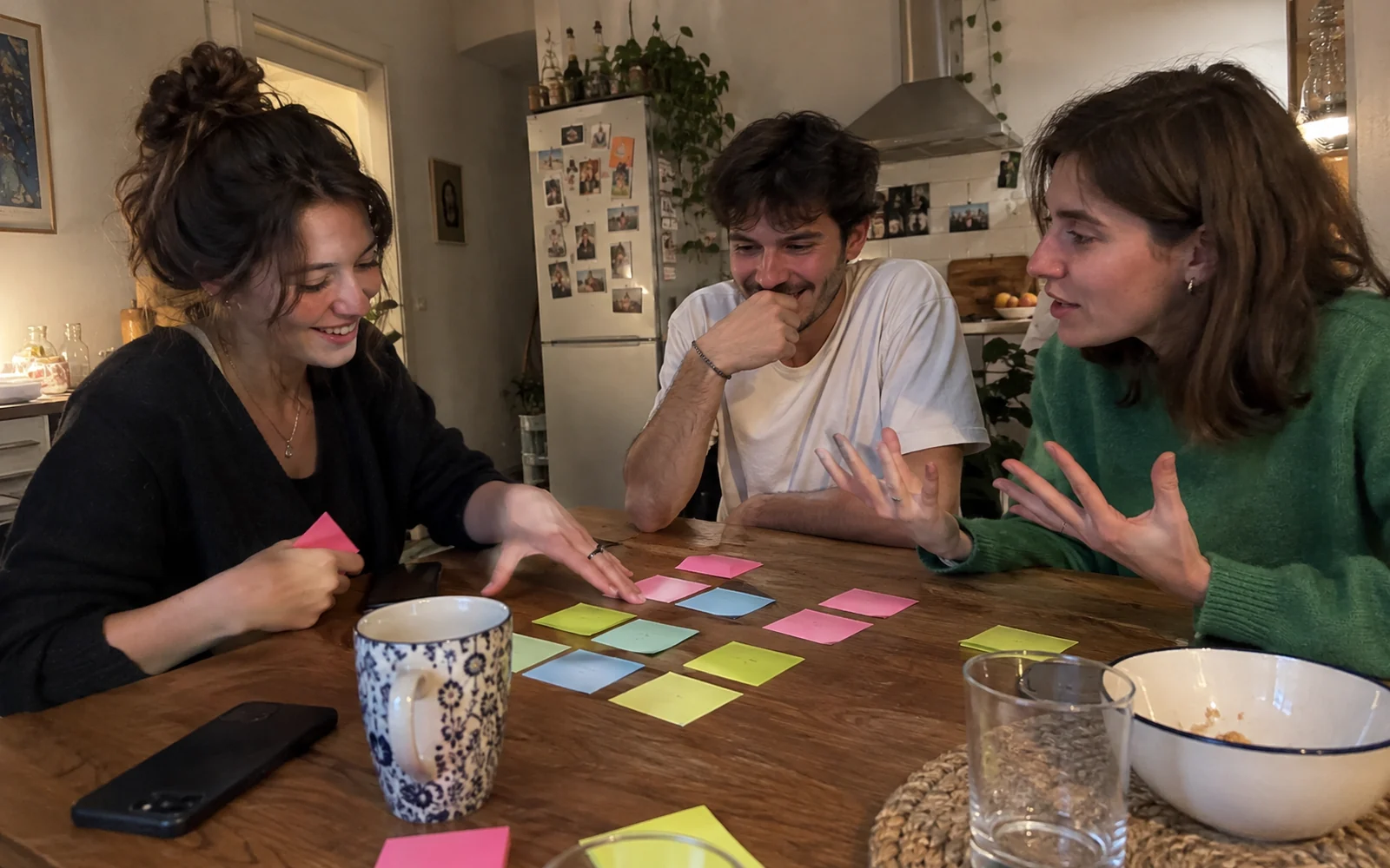 Trois amis débattent en classant des post-it colorés sur une table de cuisine.