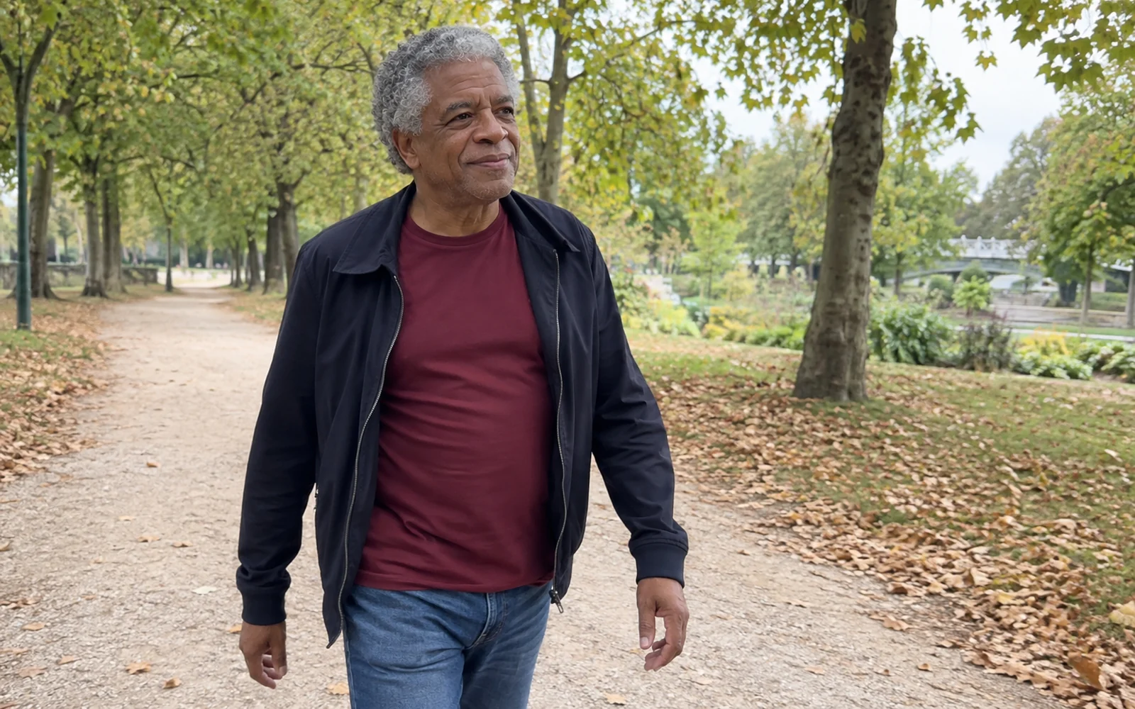 Homme aux cheveux gris et au tee-shirt bordeaux marchant dans une allée arborée