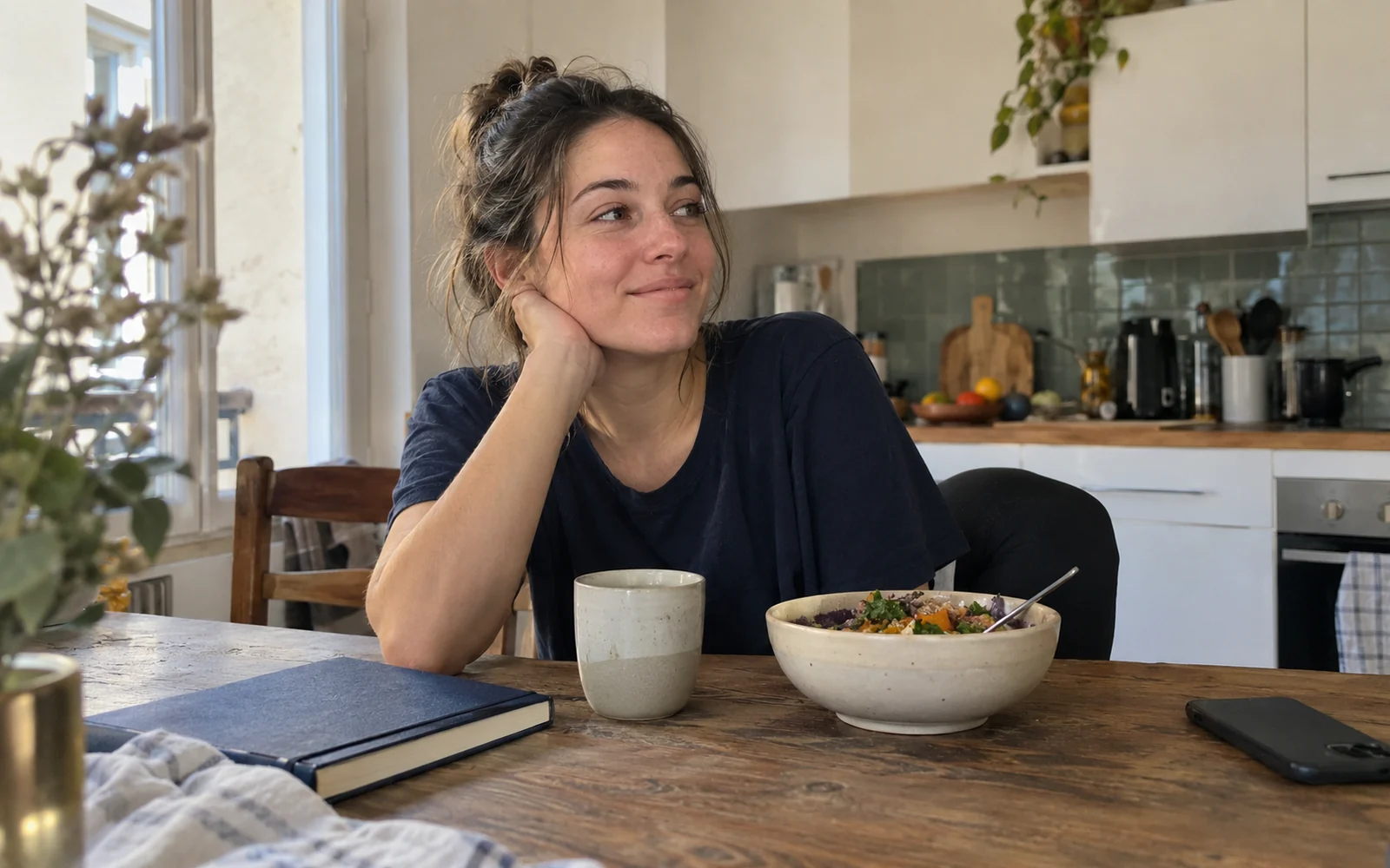 Jeune femme assise à une table de cuisine, près d’un carnet fermé, d’une tasse et d’un bol