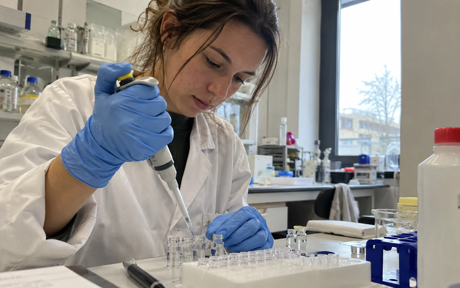 Chercheuse en blouse et gants pipetant un liquide clair dans de petits flacons de laboratoire.