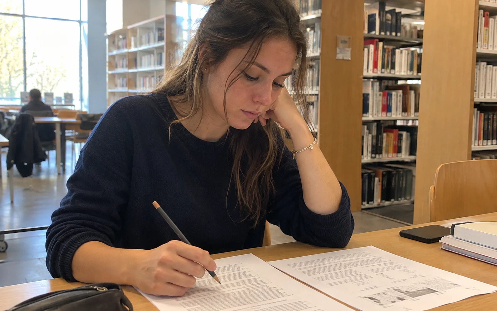 Jeune femme en pull bleu examinant des documents avec un crayon à une table de bibliothèque.