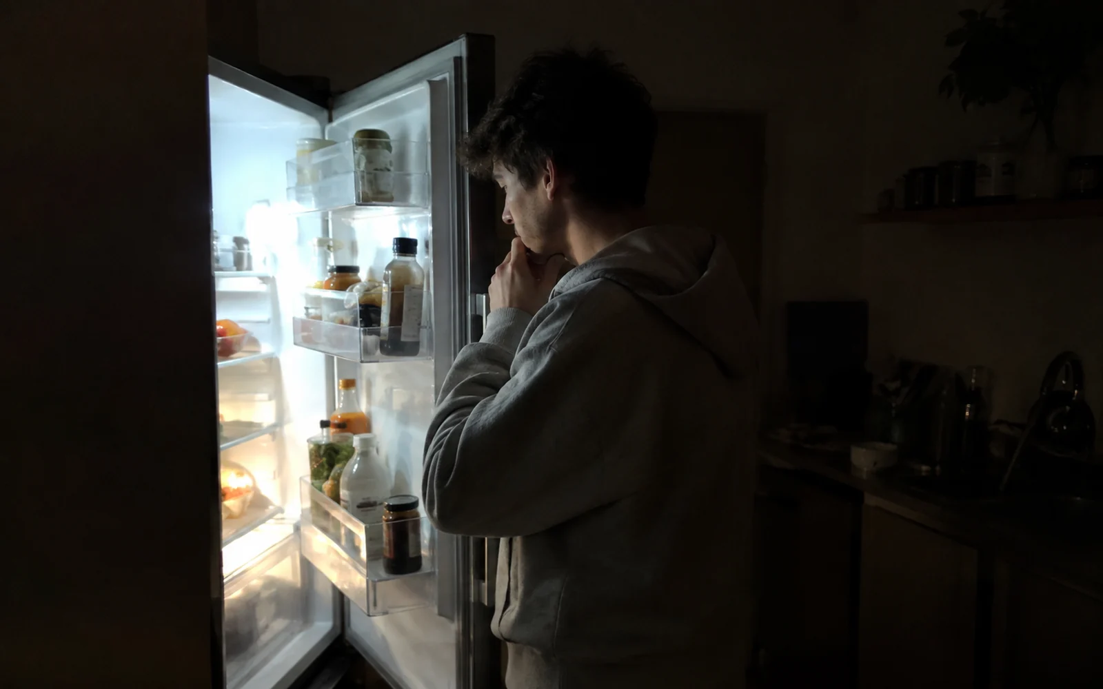 Jeune homme en sweat ouvrant le réfrigérateur la nuit, éclairé par la lumière du frigo.