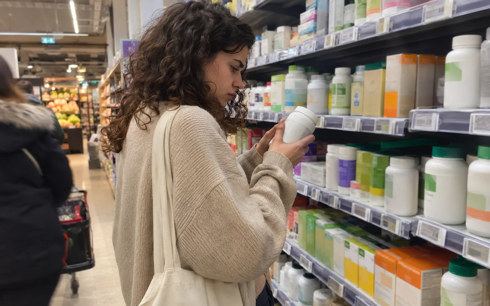 Jeune femme aux cheveux bouclés lisant le dos d’un pot de complément alimentaire dans le rayon d’un supermarché