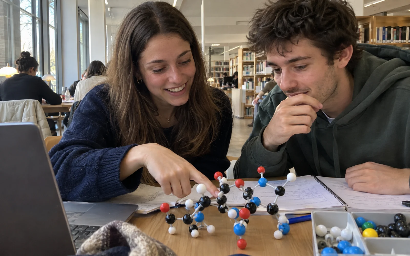 Deux étudiants observent une maquette moléculaire en plastique sur une table de bibliothèque universitaire.