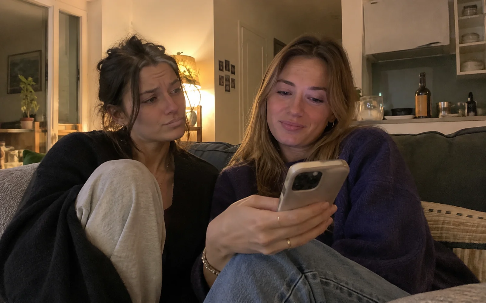 Deux amies sur un canapé regardent une vidéo sur un téléphone, l’une avec un air sceptique.