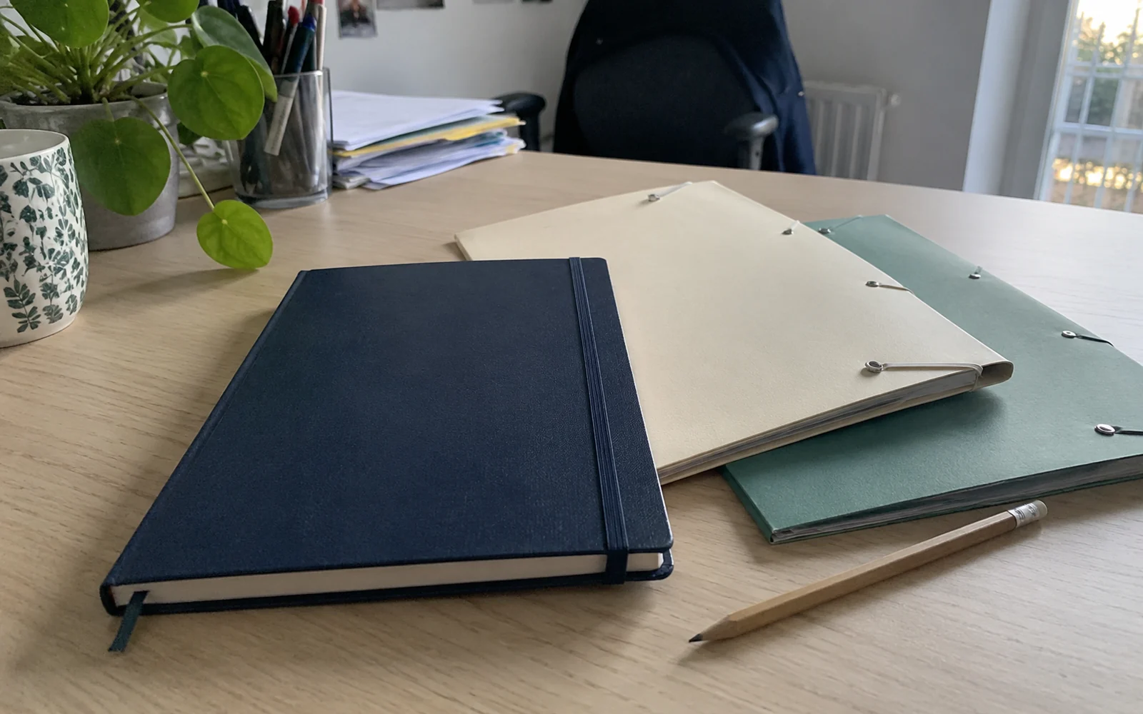 Un carnet bleu fermé, deux dossiers crème et vert et un crayon sont posés sur une table près d’une fenêtre.